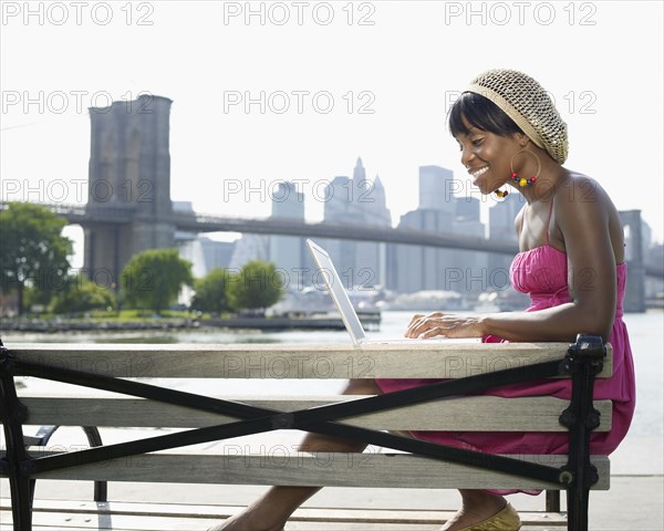 African woman sitting on bench at urban waterfront typing on laptop