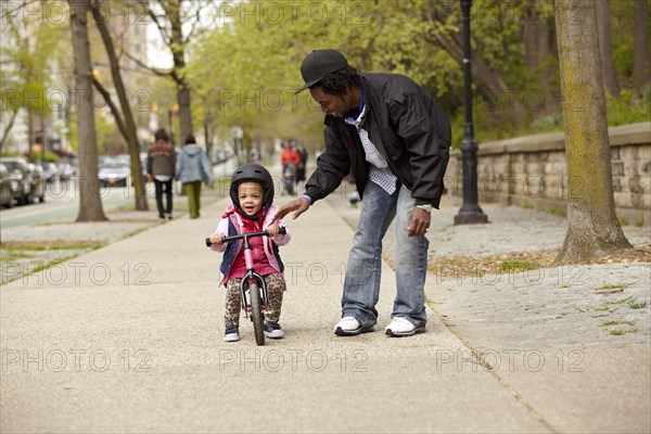 Father helping daughter ride bicycle on sidewalk