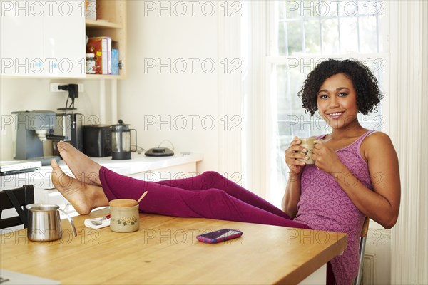 Mixed race woman drinking coffee with feet up in kitchen