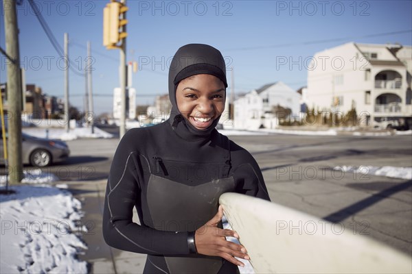 Black teenage girl in wetsuit carrying surfboard in winter