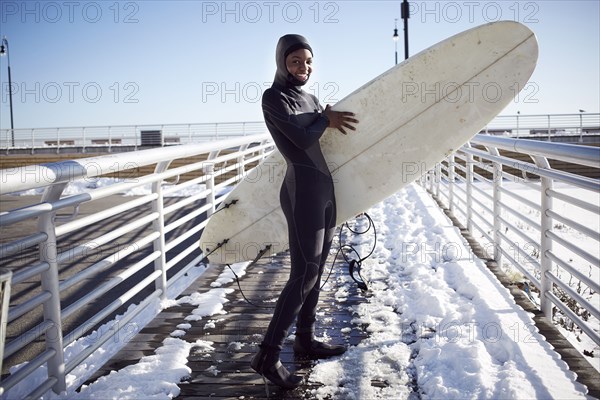 Black teenage girl in wetsuit carrying surfboard in winter