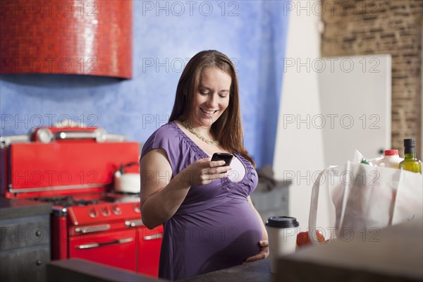 Pregnant Caucasian woman text messaging in kitchen