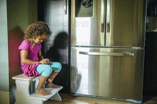 Sad mixed race girl sitting near refrigerator