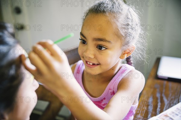 Girl painting face of mother at kitchen table