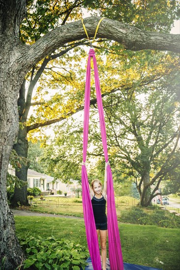 Acrobatic Caucasian girl hanging on fabric under tree