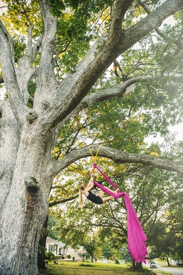 Acrobatic Caucasian girl hanging on fabric under tree