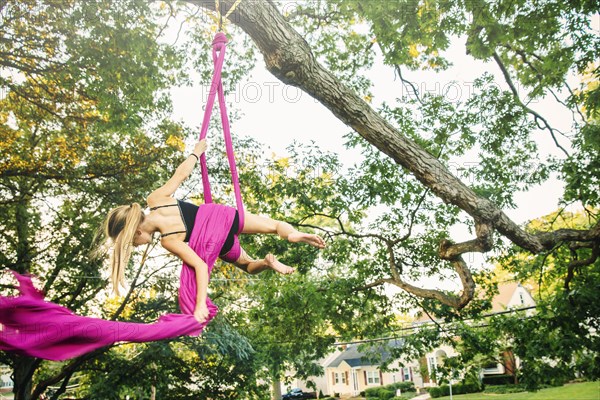 Acrobatic Caucasian girl hanging on fabric under tree