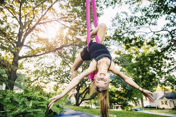 Acrobatic Caucasian girl hanging on fabric under tree