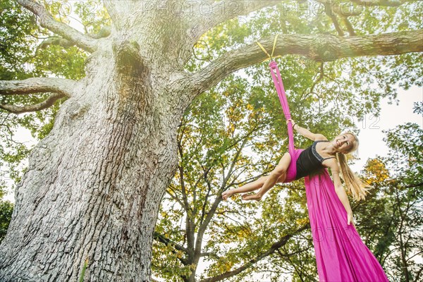 Acrobatic Caucasian girl hanging on fabric under tree