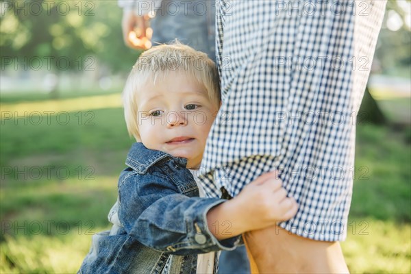 Boy clutching leg of father in grass in park