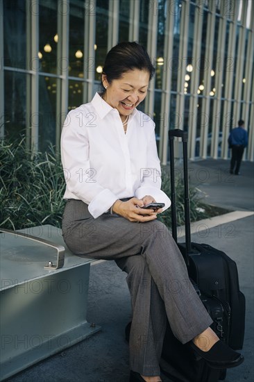 Businesswoman sitting on bench with suitcase texting on cell phone