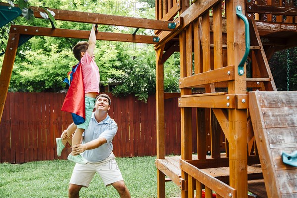 Caucasian father helping superhero son hanging on monkey bars