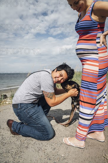 Pregnant lesbian couple playing with dog at waterfront