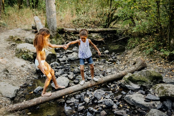 Mixed Race sisters holding hands on log over forest stream