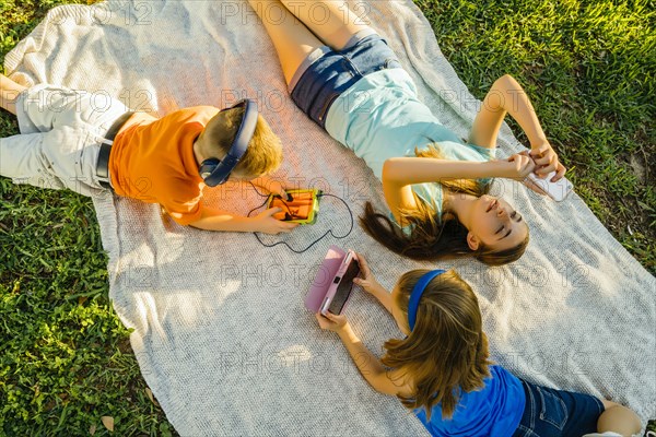 Caucasian brother and sisters laying on blanket in park using technology