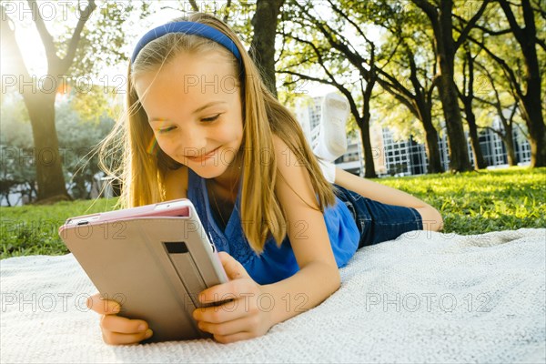 Smiling Caucasian girl laying on blanket in park reading digital tablet