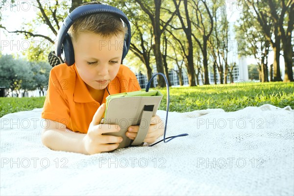 Serious Caucasian boy laying on blanket in park listening to digital tablet