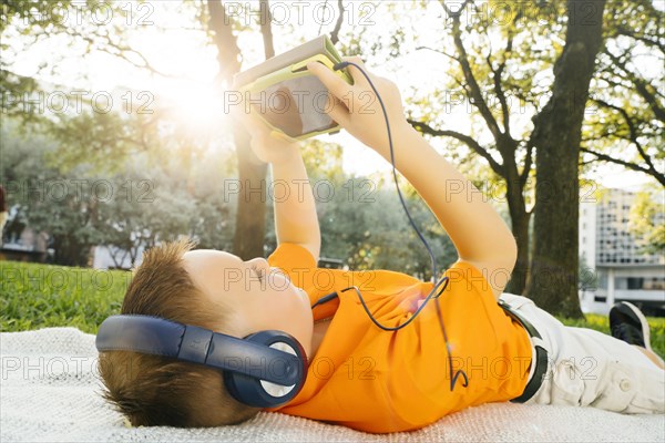 Caucasian boy laying on blanket in park listening to digital tablet