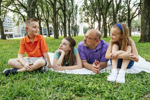 Caucasian father and daughters laying on blanket in park listening to boy