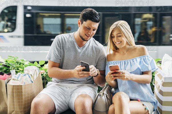 Caucasian couple with shopping bags texting on cell phones near train