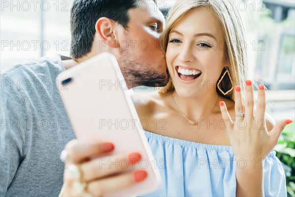 Caucasian couple holding hands and posing for selfie with engagement ring