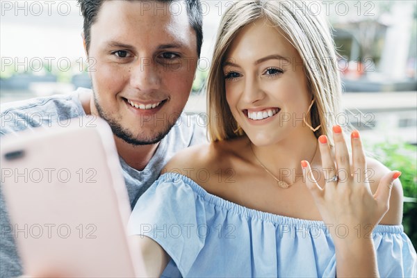 Caucasian couple posing for cell phone selfie showing engagement ring