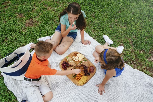 Caucasian brother and sisters eating snacks on picnic blanket