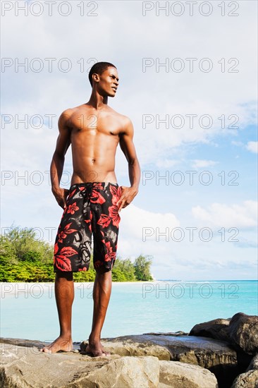 African man in swim trunks standing on rocks by ocean