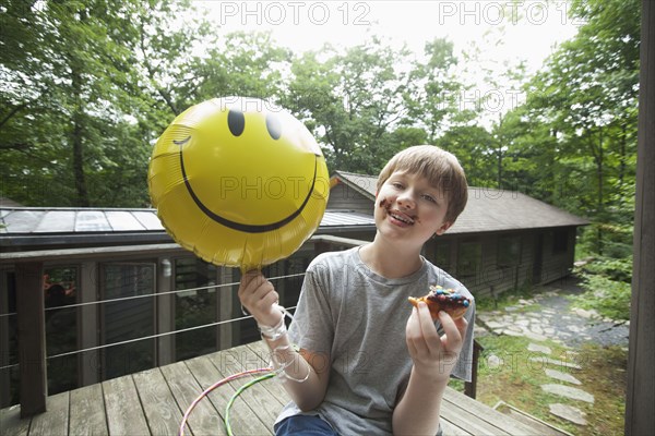 Caucasian boy with messy face holding smiley face balloon and eating donut