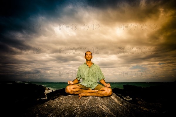 Man meditating on rock near ocean under clouds