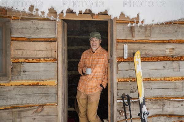 Mixed race man drinking coffee in log cabin doorway in snow