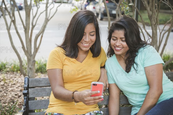 Hispanic mother and daughter using cell phone on park bench