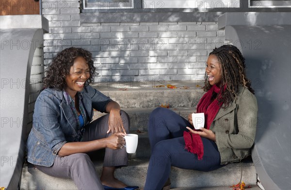 Black mother and daughter sitting on front stoop drinking coffee