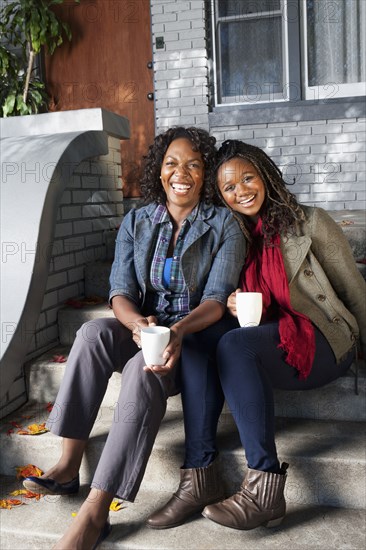 Black mother and daughter sitting on front stoop drinking coffee