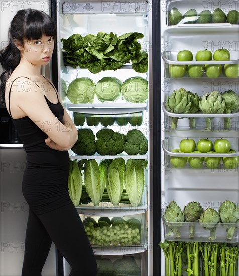 Unhappy mixed race woman looking at green vegetables in refrigerator