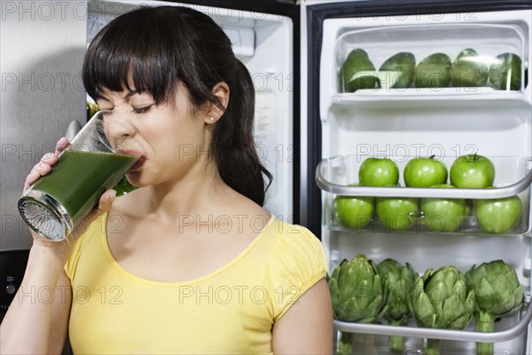 Grimacing mixed race woman drinking healthy drink near refrigerator