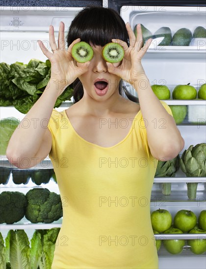 Mixed race woman covering eyes with kiwi slices near refrigerator