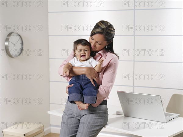 Hispanic working mother holding baby son in home office