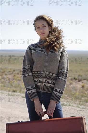 Mixed race woman standing on dirt road with suitcase