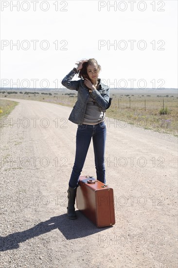 Mixed race woman standing on dirt road with suitcase