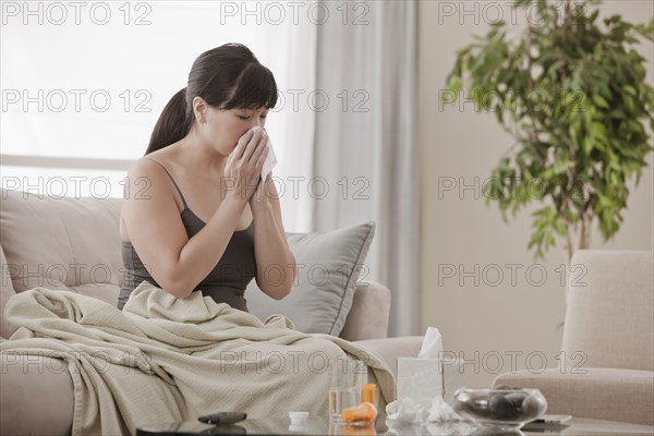 Sick Asian woman sitting on sofa blowing her nose