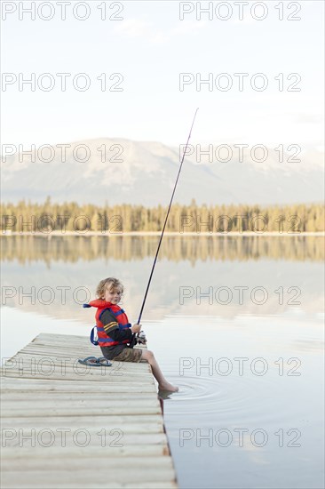 Caucasian boy sitting on dock at lake holding fishing rod