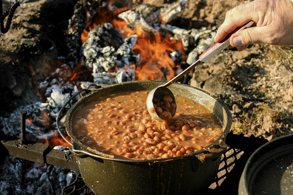 Hand of man stirring beans with ladle on campfire