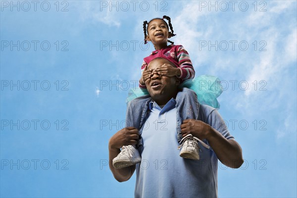 African father carrying daughter on shoulders while daughter covers his eyes