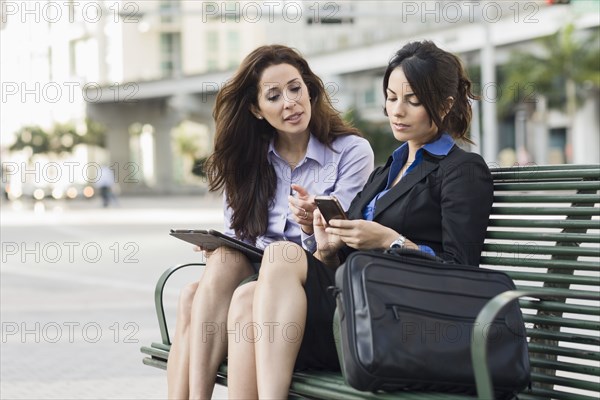 Hispanic businesswomen on bench using cell phone and digital tablet