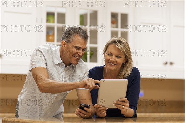 Caucasian couple using digital tablet and cell phone at kitchen counter