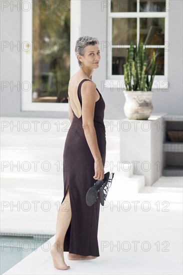 Woman in evening gown carrying shoes by swimming pool