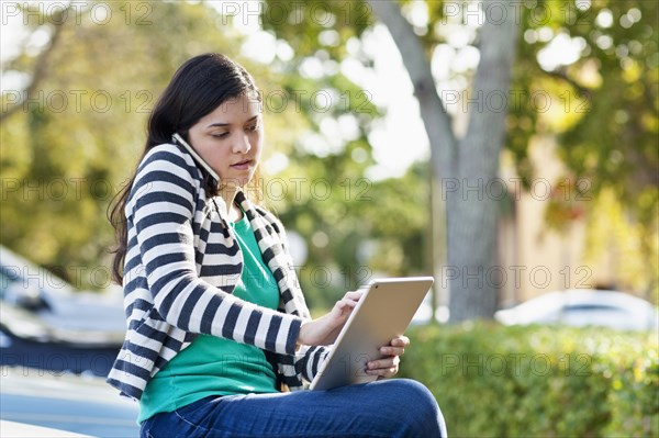 Hispanic woman using cell phone and digital tablet in parking lot