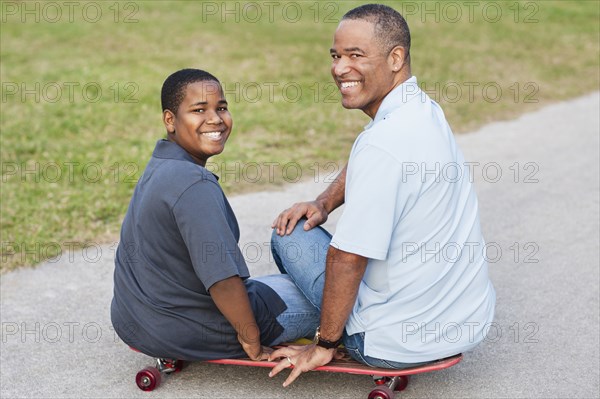 African American father and son sitting on skateboard on path