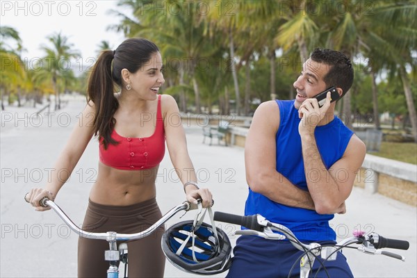 Hispanic man on bicycle talking on cell phone while girlfriend waits